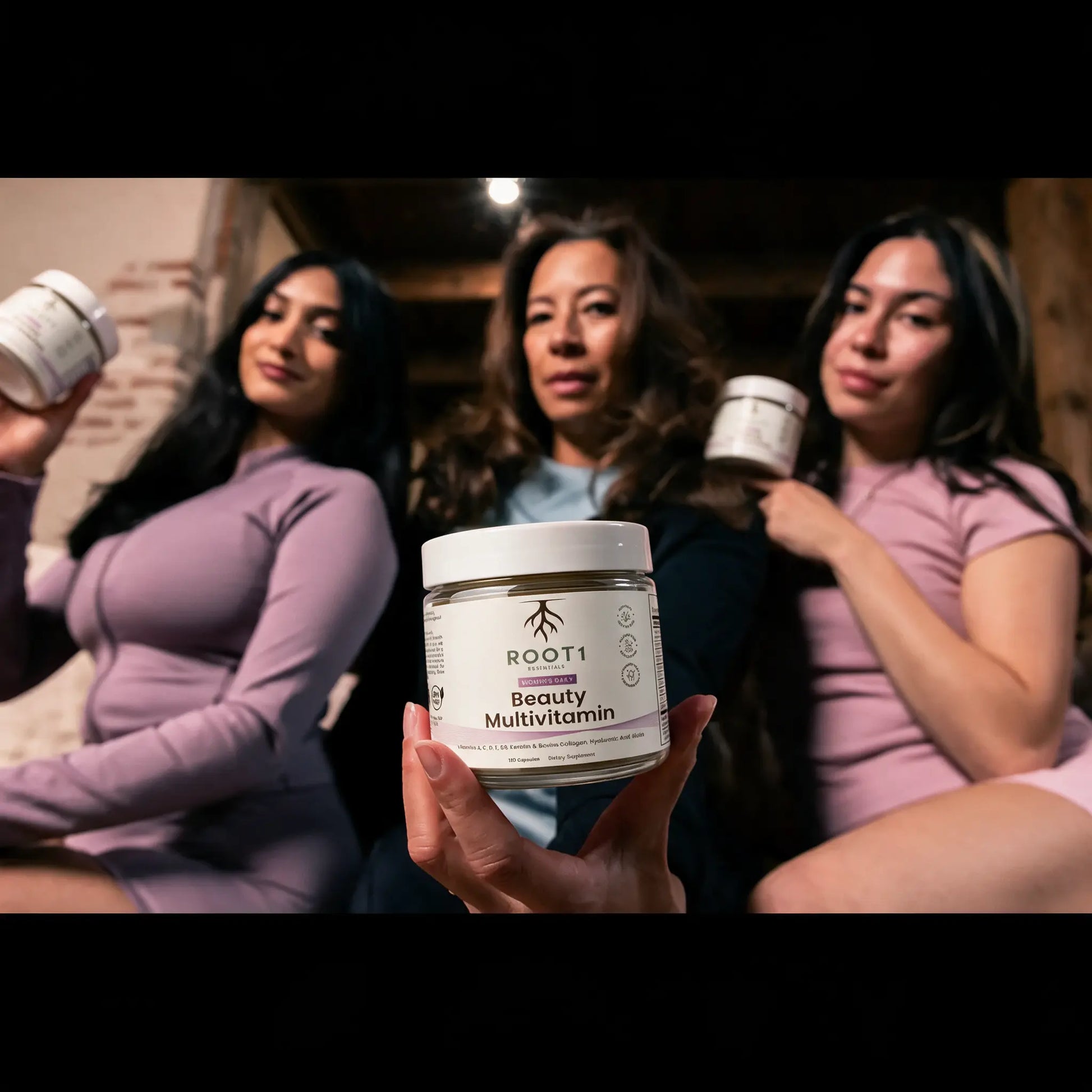 Three women are shown holding white jars of ROOT1 Beauty Multivitamin in a warm indoor setting, with one woman in the foreground holding a jar that is in sharp focus.